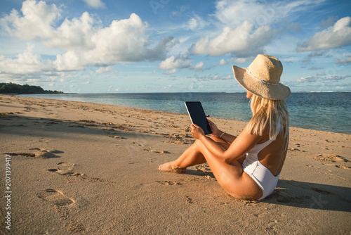Obraz na plátně Woman sits on warm sand reading a tablet near the sea