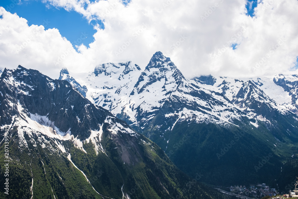 Fototapeta premium Snow-covered mountain peaks rise against a blue sky with clouds, surrounded by green valleys and rocky slopes, creating a stunning natural landscape