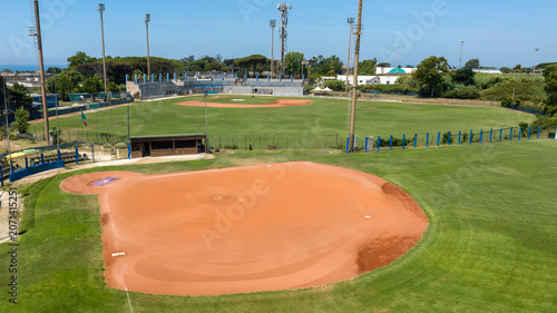 Konstfotografi Wide aerial view of a professional baseball stadium under a clear blue sky, showing the green grass outfield, detailed red dirt infield, dugouts, tall stadium light poles, and empty spectator stands