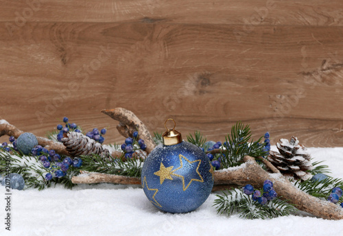 Foto Weihnachtlicher Hintergrund im Schnee mit einer blauen Christbaumkugel, Tannenzweigen und Beeren vor einem braunen Holzhintergrund mit viel Platz für Text und eigene Weihnachtsgrüße