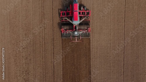 Aerial view of tractor and large multi row seed drill moving down a rectangular field, leaving darker tilled strips, radial hopper pattern, and parallel tire tracks.