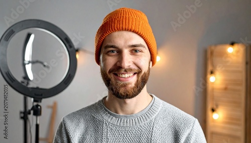 A smiling young man wearing an orange beanie hat posing in front of a ring light in a professional studio environment for video content.