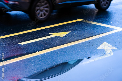 Wet asphalt road surface with yellow and white arrow markings and car reflection in puddle