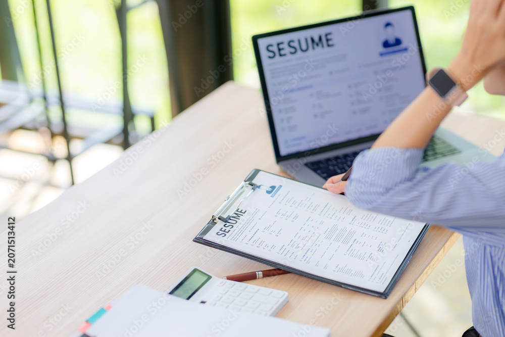Obraz premium Close up of an Asian female HR manager reviewing and examining job applicant resumes on a clipboard and laptop screen, background checking and recruitment concept.