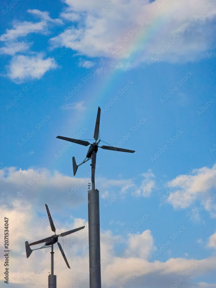 Naklejka premium Two small wind turbines generate renewable energy against a cloudy blue sky with a colorful rainbow.