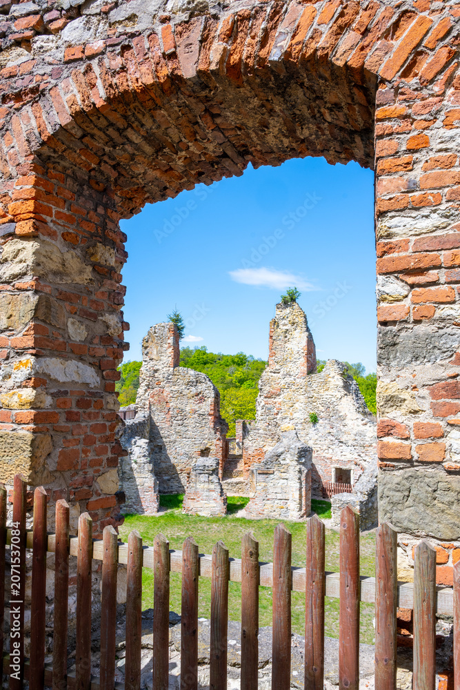 Fototapeta premium Visitors look through an archway at the ruins of Boskovice Castle. The remains show the historical structure against a clear blue sky. Green trees surround the area, marking the landscape.