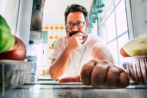 Fotografie Man standing at open refrigerator in bright kitchen, looking thoughtful while searching for food, holding chin with hand; candid domestic moment showing decision, hunger, routine and morning light