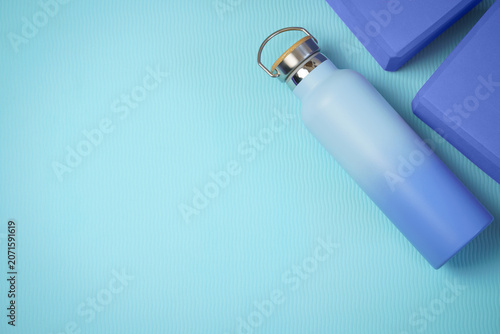A blue reusable water bottle rests beside yoga blocks on a textured mat, conveying fitness, hydration and wellness for yoga, pilates, gym workouts and eco-friendly active lifestyles.