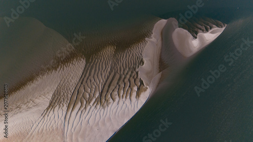 An abstract aerial perspective reveals intricate sand formations shaped by tidal forces, contrasting with deep, calm water