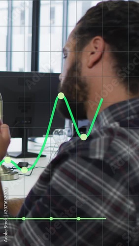 Vertical video: Man lifting cell at desk checking market as green graph peaking near chest settling