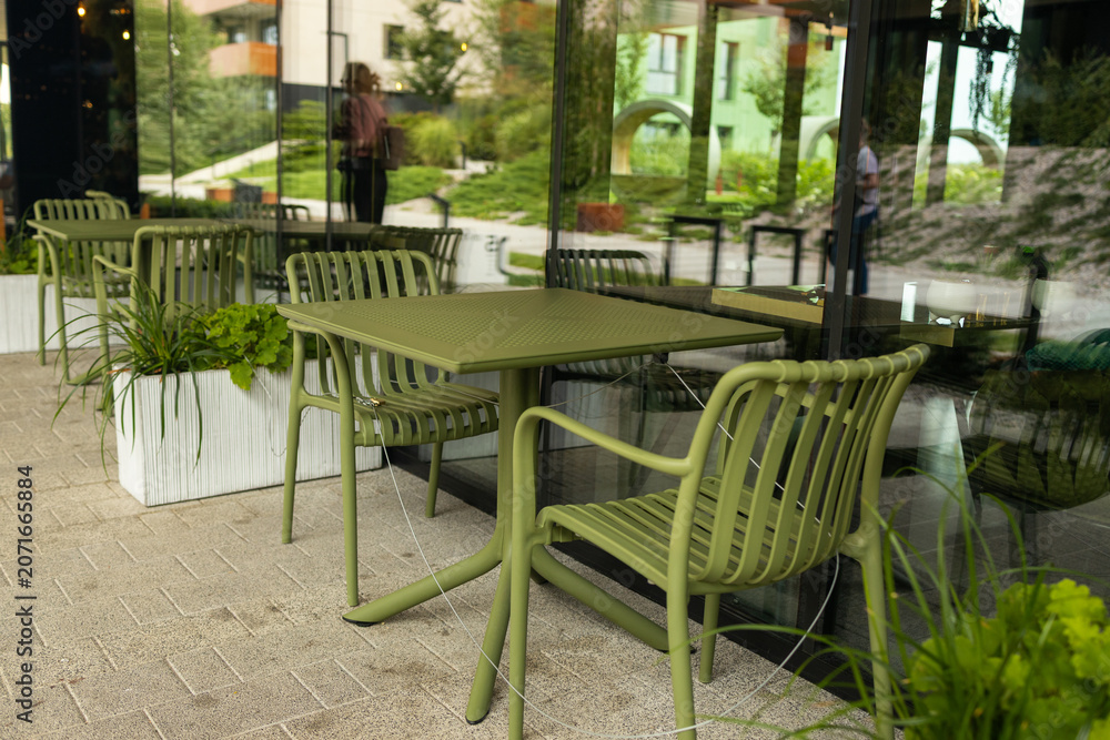 Obraz premium Stylish contemporary green metal dining table with chairs stands on an outdoor patio of a restaurant or residential courtyard, surrounded by modern landscaping and white planters.