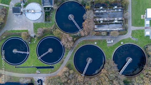 Top down aerial view of a wastewater treatment plant with circular clarifier tanks, water basins, service roads and technical infrastructure. Industrial water management facility seen from above.