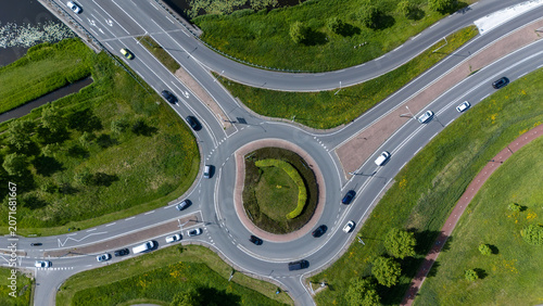 Top down aerial view of a busy roundabout with cars, curved roads, bicycle paths and green landscaping. Modern transport infrastructure and urban traffic flow seen from above.
