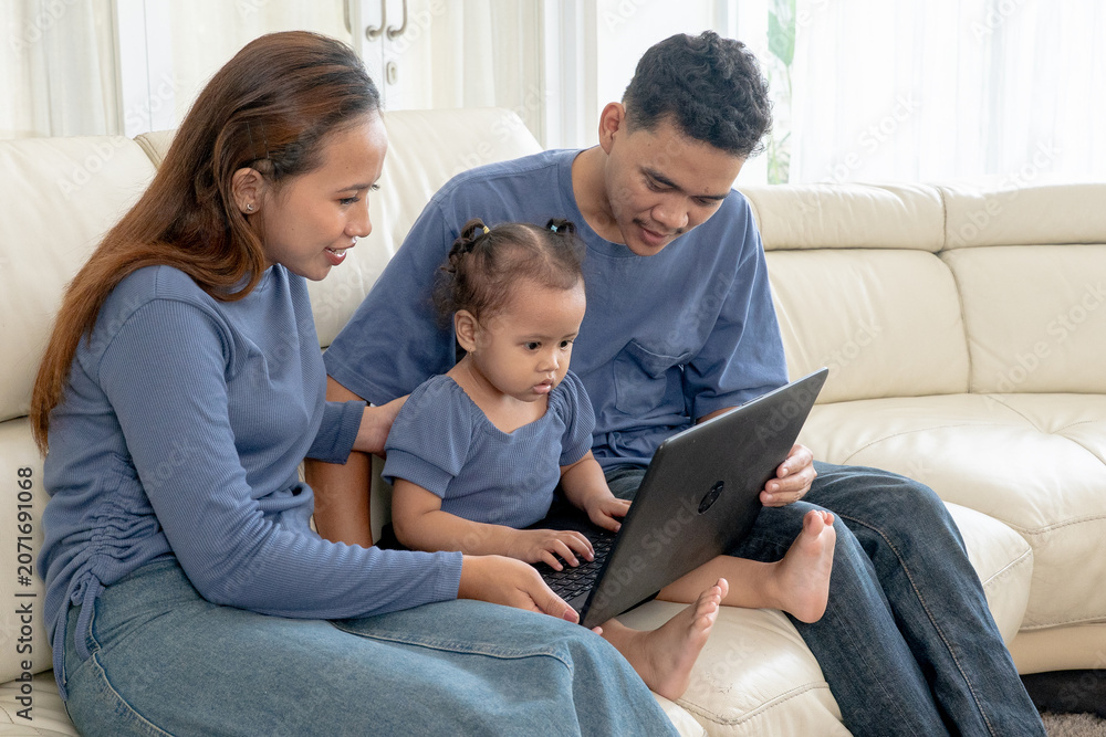 Fototapeta premium Asian Parents with Cute Toddler Daughter Watching Laptop Together on Sofa