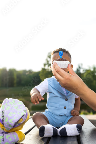 Adorable baby sitting outdoors and reaching for a birthday cupcake held by a parent. Heartwarming family celebration captured in natural evening light with a soft green background and copy space
