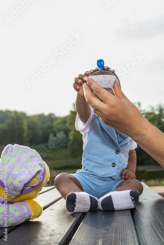 Adorable baby sitting outdoors and reaching for a birthday cupcake held by a parent. Heartwarming family celebration captured in natural evening light with a soft green background and copy space