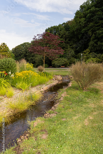 Small ornamental stream flowing through a landscaped garden surrounded by colorful flowers, shrubs and trees. Peaceful park environment with natural greenery and tranquil outdoor atmosphere