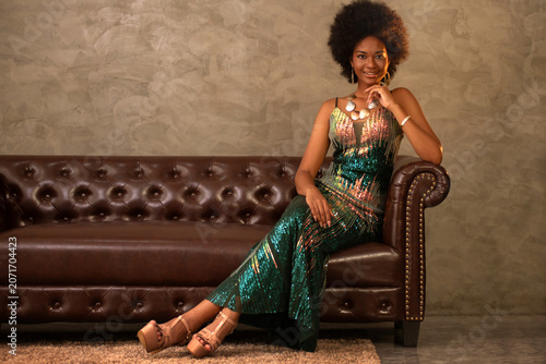 Portrait of an elegant African woman with an Afro hairstyle, wearing a long dress, sitting on a vintage-style sofa.