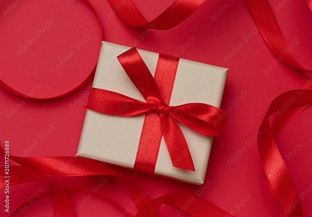Fototapeta premium Gift box in beige packaging with red satin bow on a red background