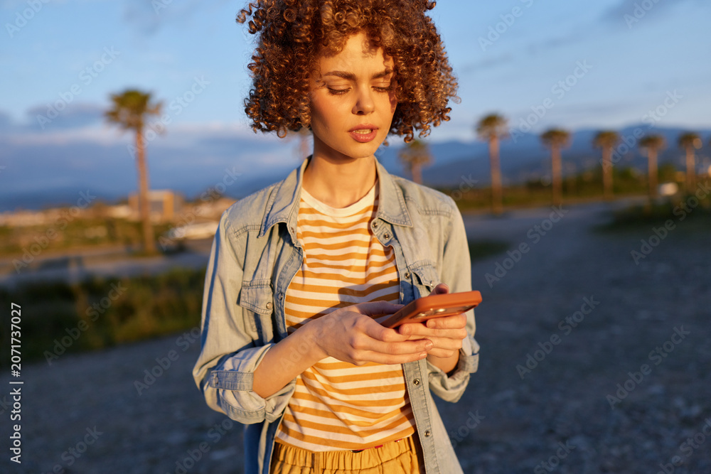 Fototapeta premium A woman in a denim jacket and striped shirt stands outdoors at golden hour, focused on her smartphone. A breezy coastal backdrop highlights casual, stylish everyday life.