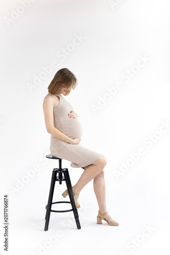 Smiling pregnant woman in beige dress sitting on stool, holding her belly.