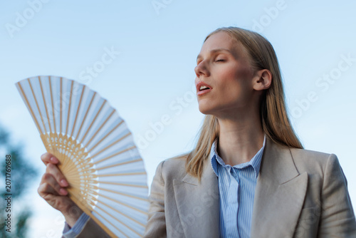 Young businesswoman uses a fan to cool down during a heatwave while outdoors on a sunny day