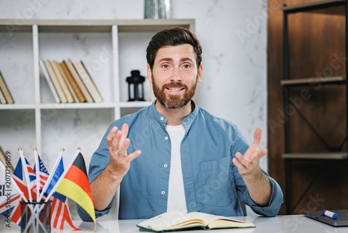 Bearded male tutor sitting at table with different flags and looking at camera