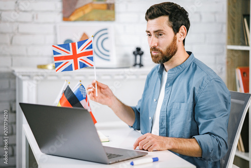 Bearded male tutor sitting at table with different flags and looking at laptop