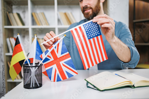 Bearded male tutor sitting on table with different flags and choose some