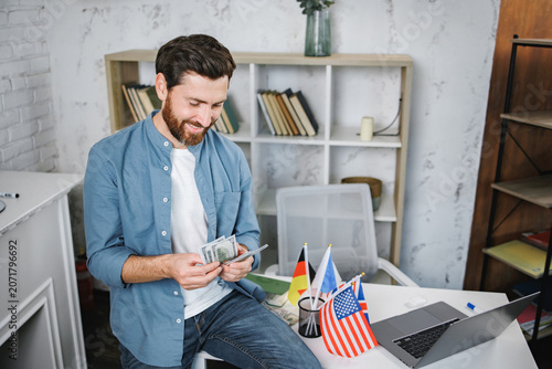 Bearded male tutor sitting on table with different flags and counting money