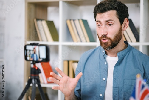 Bearded male tutor sitting at table with flags and recording a video