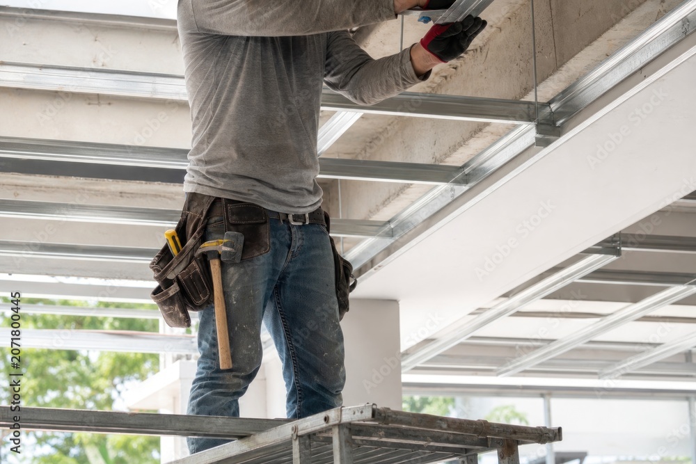 Fototapeta premium Skilled construction worker installing metal ceiling frame structure on scaffolding inside modern building site, professional contractor wearing tool belt working on active renovation project.