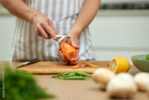 Hands peeling a carrot with a peeler on a wooden cutting board, preparing healthy fresh vegetables in a home kitchen