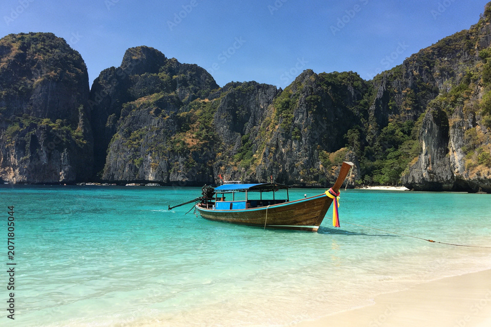 Obraz premium Thai longtail boat floats on turquoise sea beside white sandy beach, towering limestone cliffs under clear blue sky.