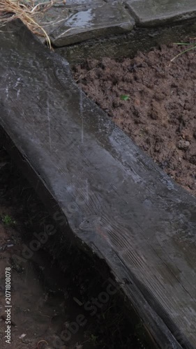 Rainwater flows on a wooden plank near wet soil in an open area during a light rainfall in a rural setting