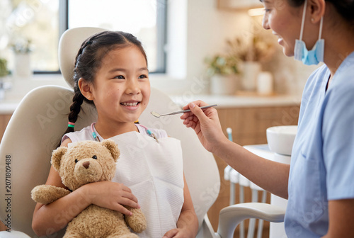 A cheerful young girl with braided hair hugs her teddy bear while smiling at a friendly dentist holding a small mirror nearby