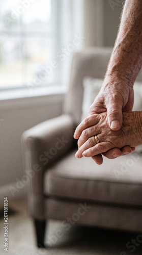 Close up of wrinkled elderly hands tenderly holding each other with a gold wedding ring visible in a blurred cozy living room