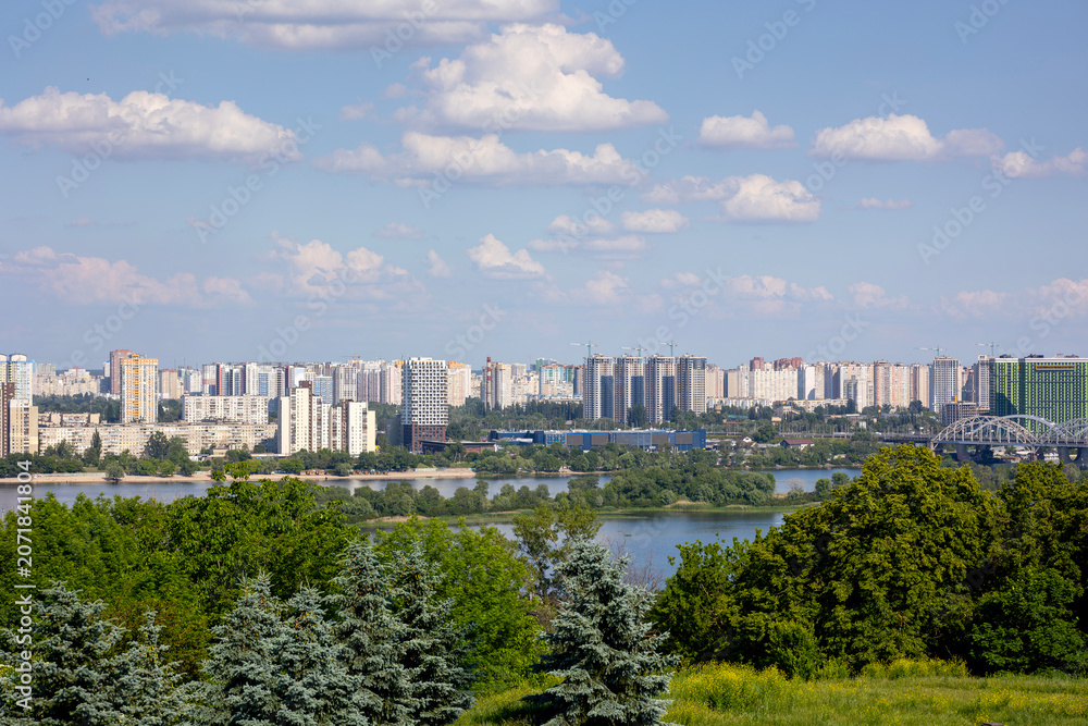 Fototapeta premium Kyiv, Ukraine, a panoramic view across the Dnipro River, revealing the city's left bank residential areas, modern high-rise buildings, and the Darnytskyi railway bridge under a clear sky with clouds