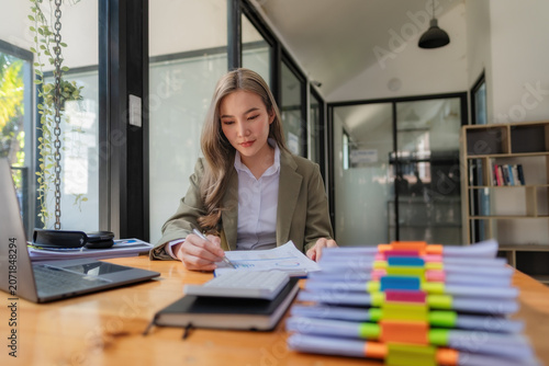 Asian businesswoman writing and checking information on paperwork with a large stack of documents on office desk.