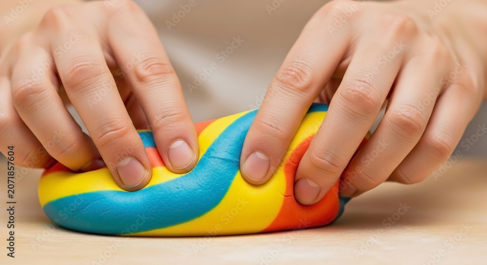 Fototapeta premium Close up of hands kneading colorful striped play dough. Young person molding bright red yellow and blue clay on a wooden table. Creative tactile activity