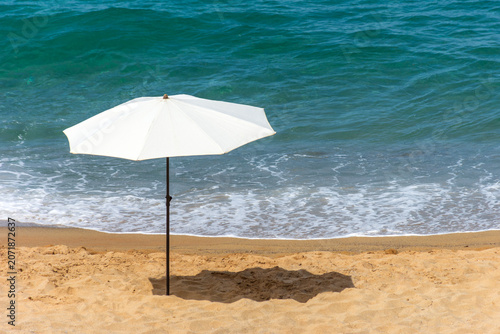 White beach umbrella on sandy shore with gentle blue Mediterranean waves.