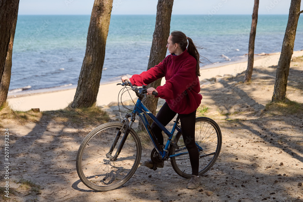 Fototapeta premium Young woman with brown hair riding blue mountain bike on sandy beach path near ocean under sunlight