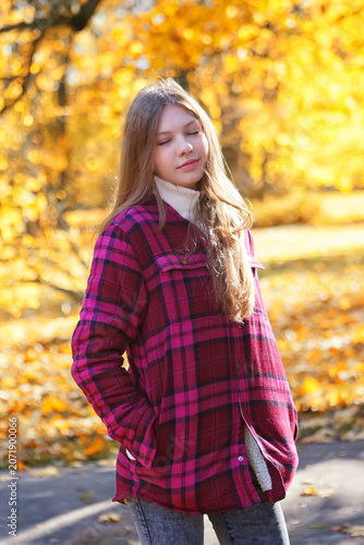 Girl stands in a park surrounded by yellow leaves during autumn while wearing a flannel shirt and looking relaxed