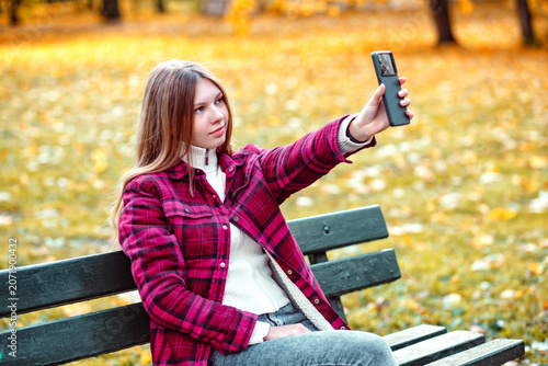 Young woman takes selfie with smartphone while sitting on bench in park during autumn season