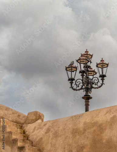 Traditional wrought iron street lamp on a clay wall with pigeons under a cloudy sky in the old Medina of Marrakech, Morocco