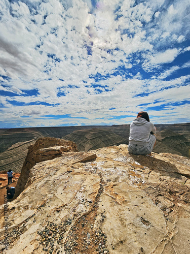 Goosenecks State Park reserve, Utah, USA. Young woman, a tourist, admiring the canyons and reddish meanders sculpt by the San Juan River over centuries against a blue cloudy sky. UNESCO heritage site.
