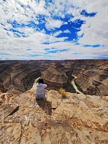 Goosenecks State Park reserve, Utah, USA. Young woman, a tourist, admiring the canyons and reddish meanders sculpt by the San Juan River over centuries against a blue cloudy sky. UNESCO heritage site.