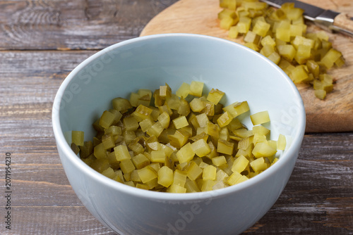 Finely diced pickles in a bowl with chopped cucumber pieces on a cutting board, prepared as an ingredient for cooking on a rustic wooden surface