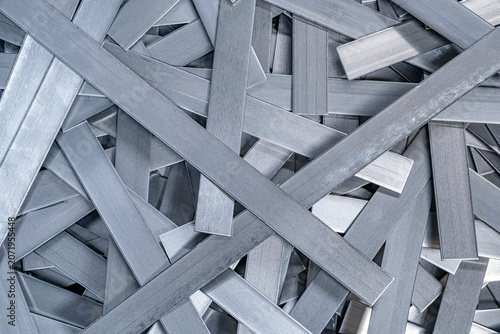 Overlapping flat metal strips in a dense pile. The brushed steel pieces create a complex industrial texture with a modern, professional and cold monochromatic vibe. Modern industrial manufacturing.