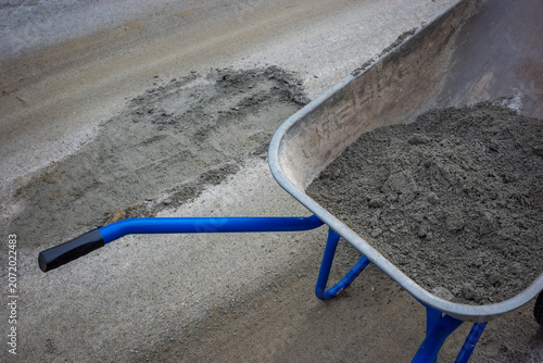 Metal wheelbarrow with cement mixture near patched asphalt pothole on suburban road. Concept: road repair